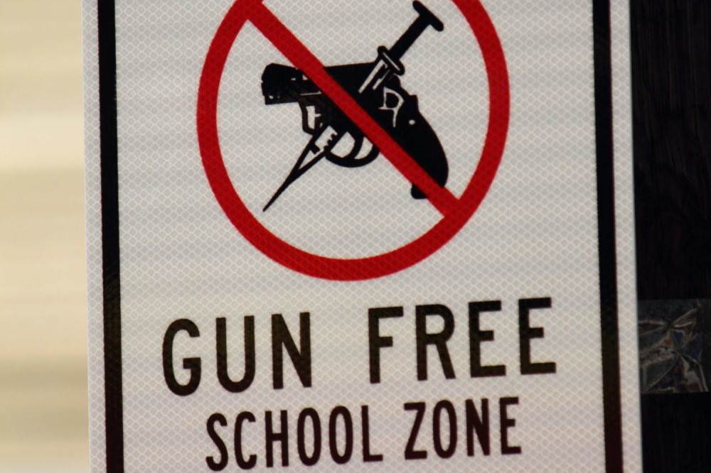 White road sign with bold black capital letters stating, “Drug Free Gun Free School Zone”. The smaller print states, “Violators will face severe federal, state, and local criminal penalties.” There is also an image of a gun with a red circle and slash overtop signifying a Gun Free School Zone.